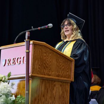 Cassandra Colon delivers her graduate reflection wearing academic regalia at a Regis branded podium