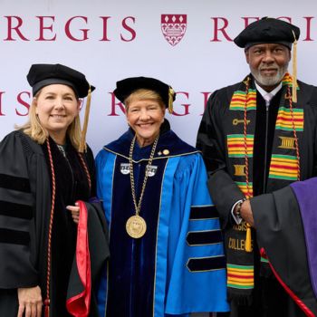 Charlotte Cramer Wagner, President Antoinette Hays, and Alix Cantave wearing academic regalia in front of a Regis backdrop