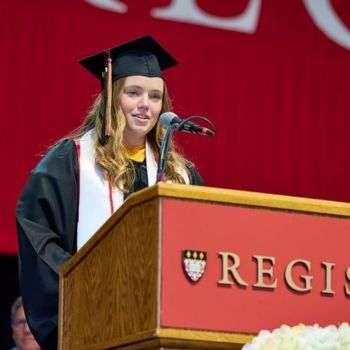 Madeline Conover delivers her graduate reflection wearing academic regalia at a Regis branded podium