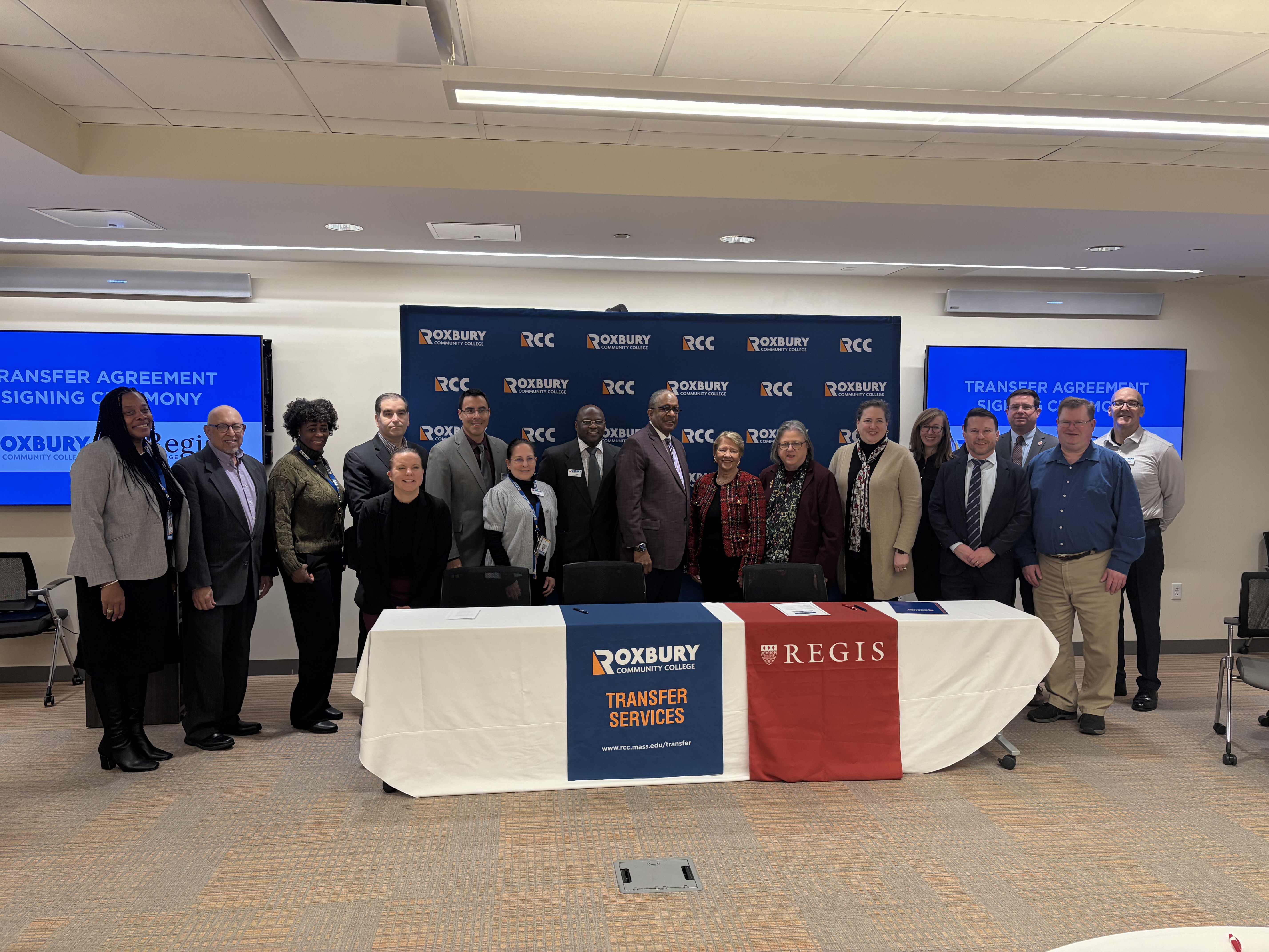 Seventeen members of staff and faculty from Regis College and Roxbury Community stand behind a table after signing an agreement.